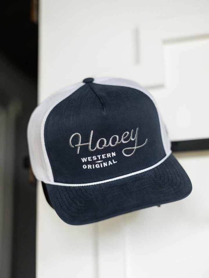 A navy blue and white Hooey Colby Cap by Savannah Sevens western life{&}style with "Western Original" embroidery hangs on a white door, featuring an adjustable snap back for comfort.