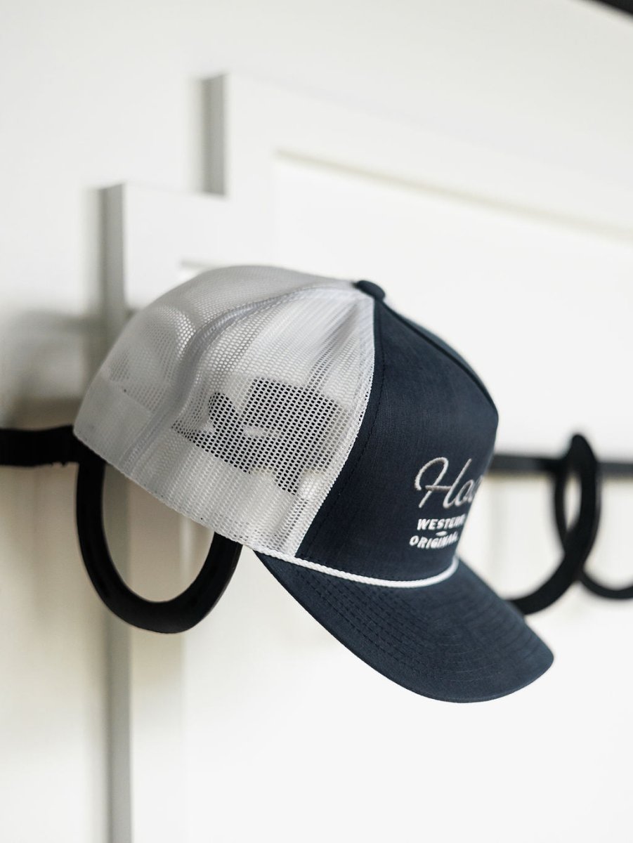 The Hooey Colby Cap by Savannah Sevens western life{&}style is a black and white trucker hat with white front text, hanging on a black hook against a white wall. It features an adjustable snap back for the perfect fit.