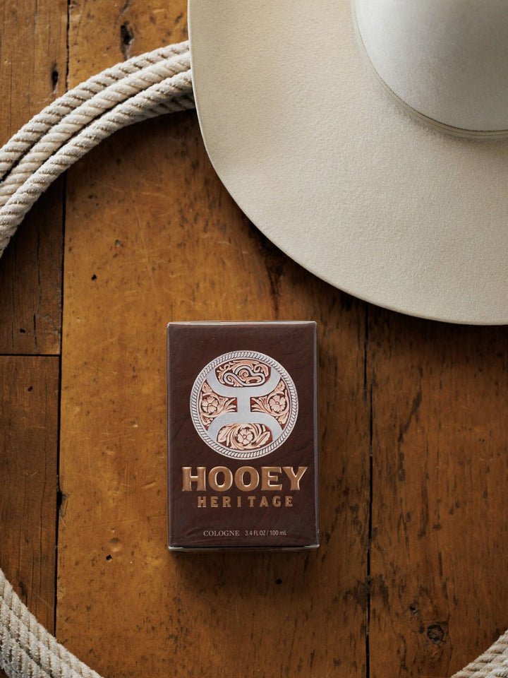 A Hooey Heritage Cologne by Savannah Sevens western life{&}style sits on a wooden surface beside a coiled rope and cowboy hat, capturing the essence of cowboy cologne with its subtle leather scent.