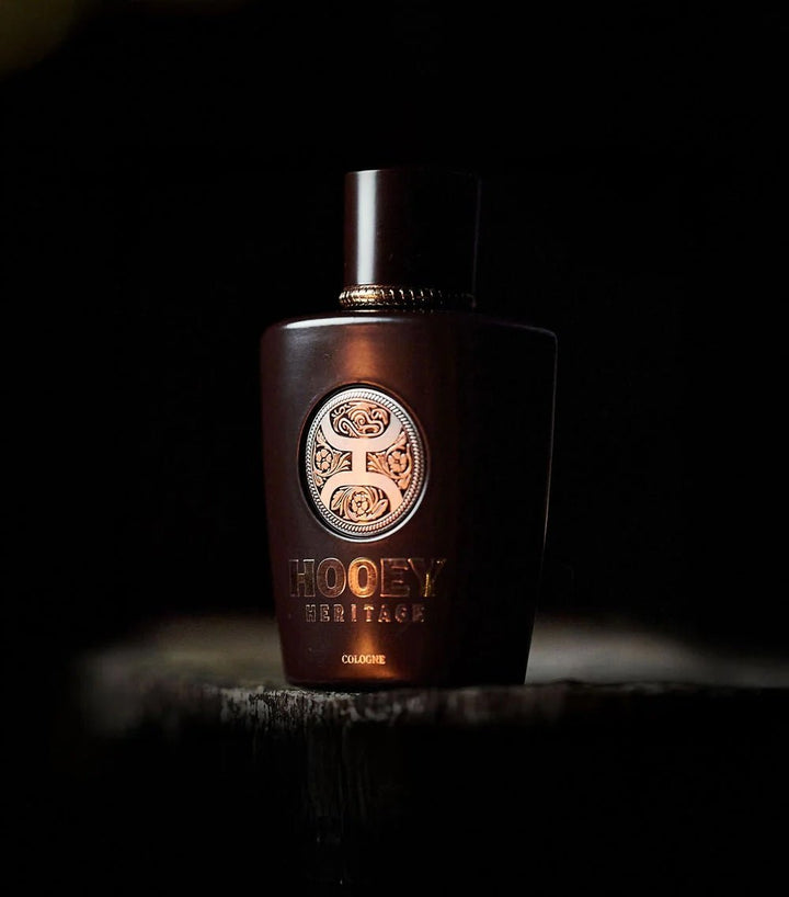A dark brown bottle of Hooey Heritage Cologne by Savannah Sevens western life{&}style, featuring a gold embossed logo, sits on textured wood against a dark background, evoking the scent of rich leather.