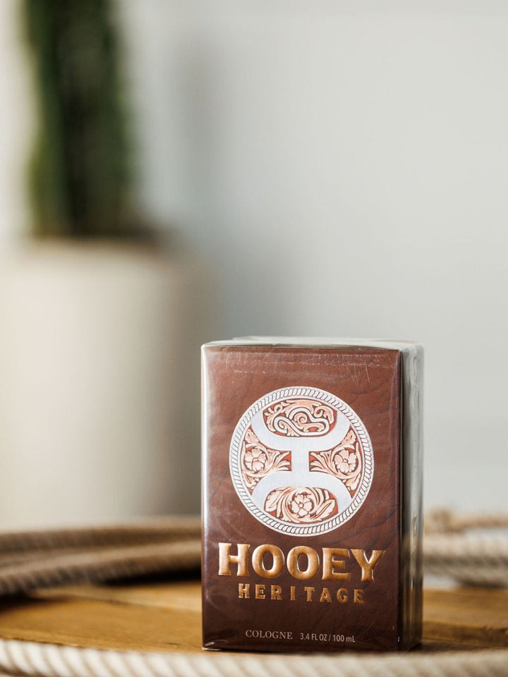 A boxed bottle of Hooey Heritage Cologne by Savannah Sevens western life{&}style sits on wood. Its brown packaging with gold lettering and a circular design hints at the rugged, leather-inspired scent, while the blurred background adds warmth.