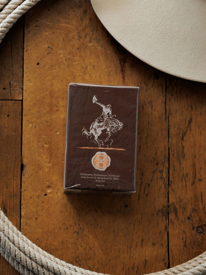 A brown box with a white cowboy illustration, beside a coiled rope and beige hat on wood, captures the rugged charm of Hooey Heritage Cologne by Savannah Sevens western life{&}style and the allure of a classic leather scent.