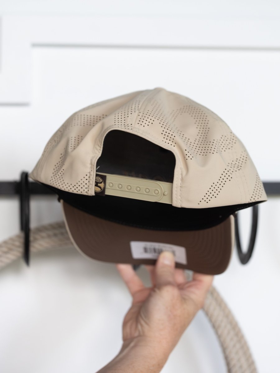A hand holds the Hooey Lansford Cap by Savannah Sevens western life{&}style, shown from the back with a perforated design, adjustable snap back, and brown underside of the brim. The background is blurred.
