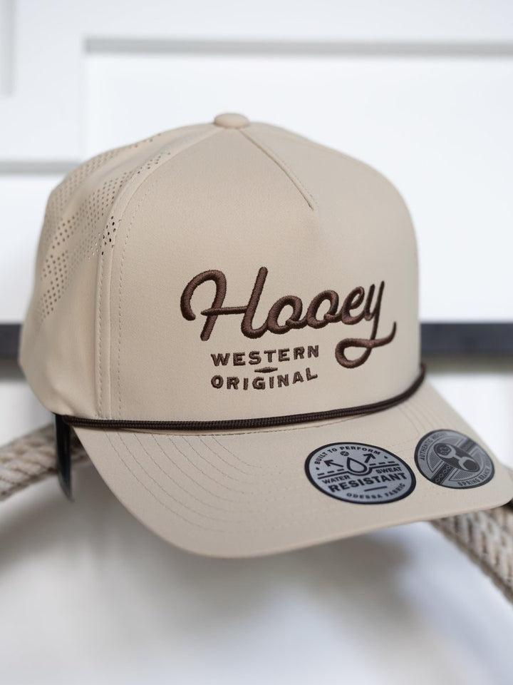 The Savannah Sevens western life{&}style Hooey Lansford Cap is a beige trucker hat with "Hooey Western Original" brown embroidery, an adjustable snapback, a rope detail above the brim, and two stickers on the flat bill.