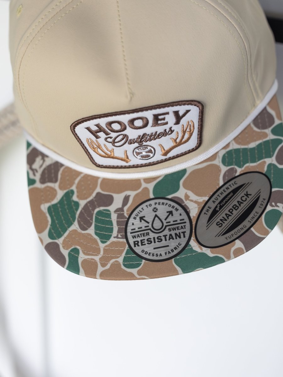The Hooey Outfitters Cap by Savannah Sevens western life{&}style features a beige crown, camouflage brim with an antlers patch, and stickers showcasing its water and sweat resistance. Crafted from durable fabric.