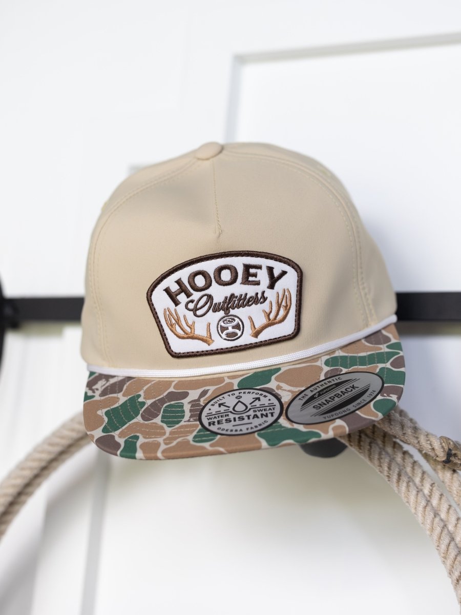 A tan Hooey Outfitters Cap by Savannah Sevens western life{&}style hangs on a hook, featuring a camo bill, brand name and antler graphics on front, plus stickers showing its water-resistant and snapback design.