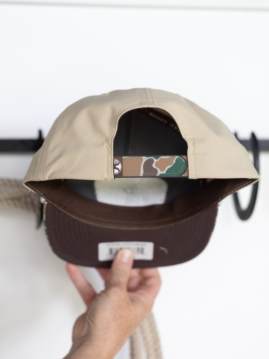 A hand holds the Savannah Sevens western life{&}style Hooey Outfitters Cap, showing its tan patch front, camo-patterned adjustable strap, and dark brown brim underside; a white background has black hooks and a coiled rope.