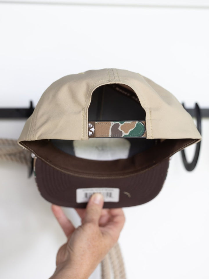 A hand holds the Savannah Sevens western life{&}style Hooey Outfitters Cap, showing its tan patch front, camo-patterned adjustable strap, and dark brown brim underside; a white background has black hooks and a coiled rope.