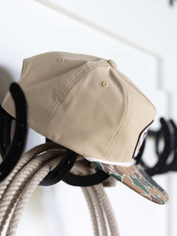 A Hooey Outfitters Cap by Savannah Sevens western life{&}style with a tan camo bill and camouflage brim hangs on a hook.