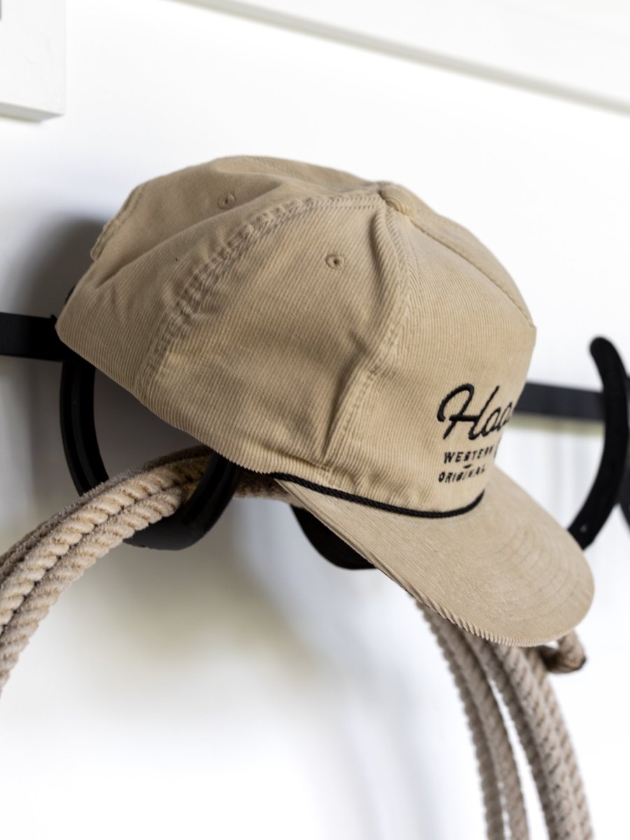 The Hooey Wylder Cap by Savannah Sevens western life{&}style features beige corduroy with black embroidery, hanging on a wall hook beside a rope—a classic western look for trucker cap fans.