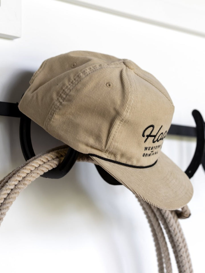 The Hooey Wylder Cap by Savannah Sevens western life{&}style features beige corduroy with black embroidery, hanging on a wall hook beside a rope—a classic western look for trucker cap fans.