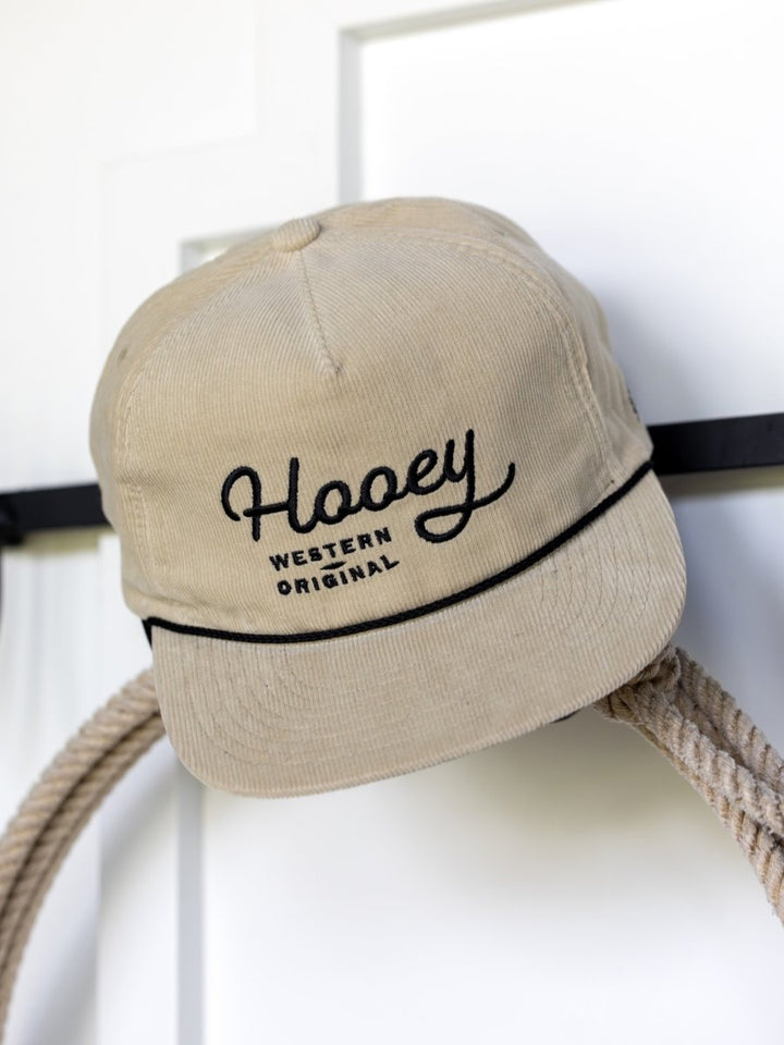 A beige Hooey Wylder Cap by Savannah Sevens western life{&}style, featuring "Western Original" in black embroidery, hangs on a hook beside a coiled rope.