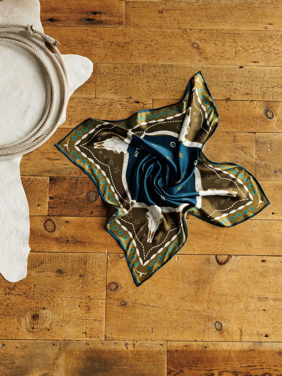 The Hooked Neck Scarf by Savannah Sevens western life{&}style, in blue, brown, and gold with white horse prints, is displayed on a wooden floor beside a white cowhide rug and a coiled rope.