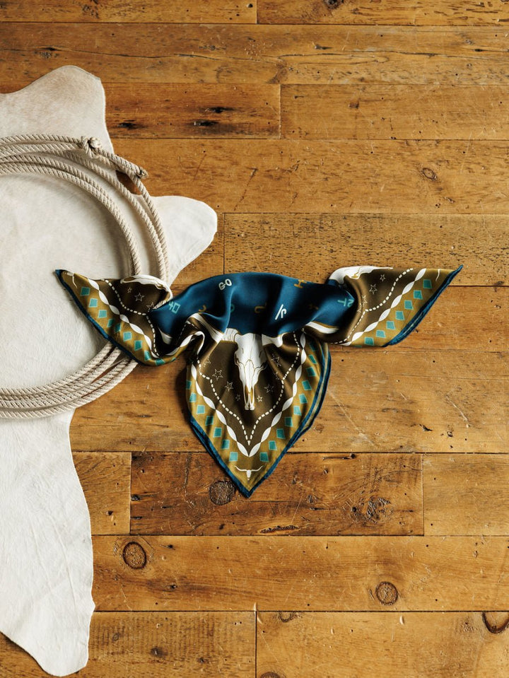 The Hooked Neck Scarf by Savannah Sevens western life{&}style, featuring a bold pattern, is laid flat on wood beside a coiled rope and white cowhide rug, showcasing classic western silk scarf style.