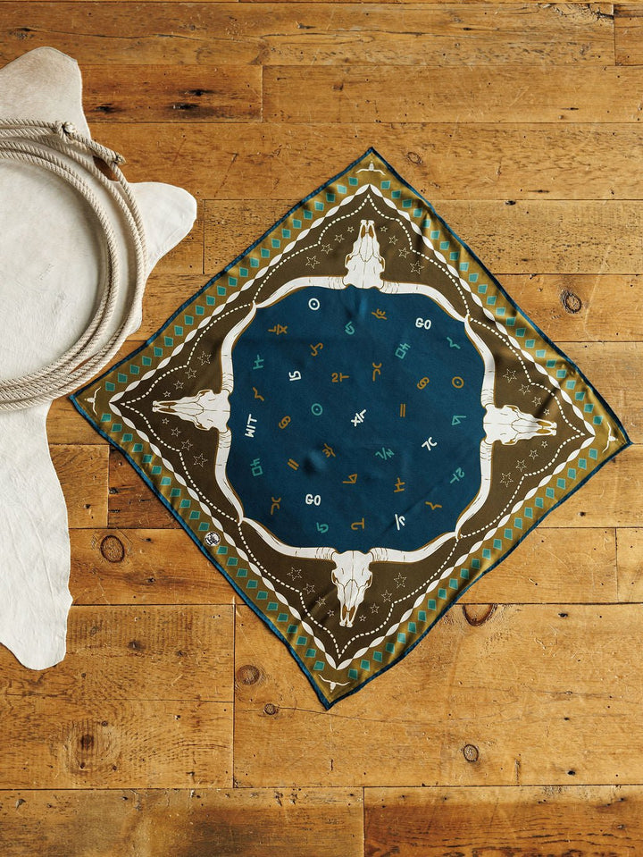 The Hooked Neck Scarf by Savannah Sevens western life{&}style is a square silk scarf with a southwestern steer skull and zodiac design in blue and olive-green, shown flat on wood beside coiled rope and a white fur rug.