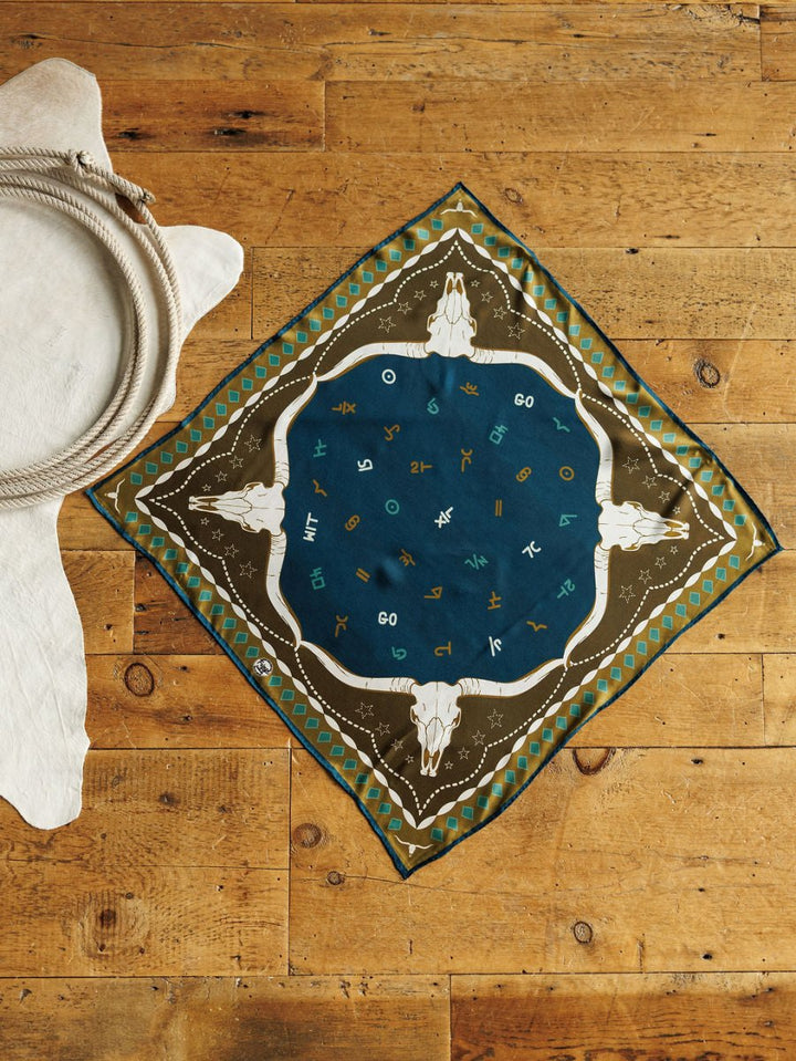 The Hooked Neck Scarf by Savannah Sevens western life{&}style is a square silk scarf with a southwestern steer skull and zodiac design in blue and olive-green, shown flat on wood beside coiled rope and a white fur rug.