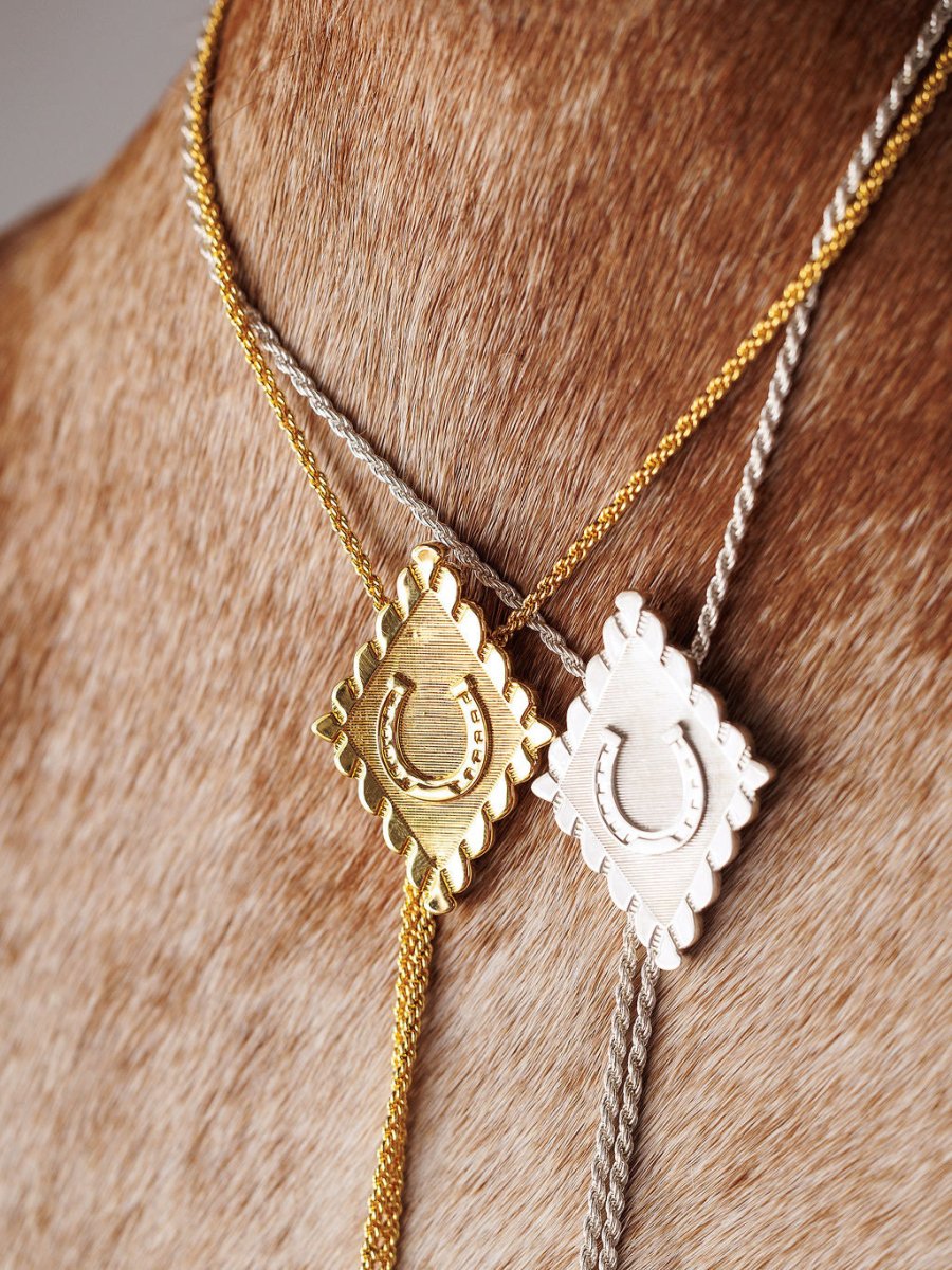 The Savannah Sevens western life{&}style Horseshoe Bolo necklaces—one gold plated and one sterling silver, both with square, scalloped-edge horseshoe pendants—are shown on a brown cowhide background.