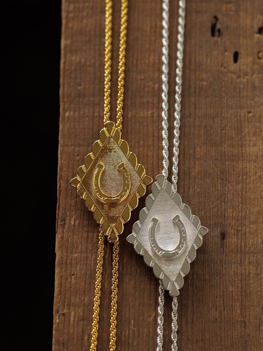 The Horseshoe Bolo necklaces by Savannah Sevens western life{&}style feature diamond-shaped horseshoe pendants with textured backgrounds and matching chains in gold plated and sterling silver, displayed on a wooden surface.