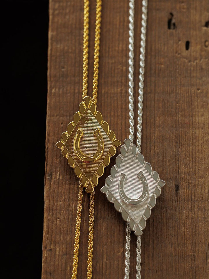 The Horseshoe Bolo necklaces by Savannah Sevens western life{&}style feature diamond-shaped horseshoe pendants with textured backgrounds and matching chains in gold plated and sterling silver, displayed on a wooden surface.