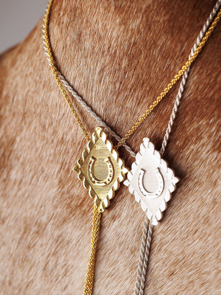 The Savannah Sevens western life{&}style Horseshoe Bolo necklaces—one gold plated and one sterling silver, both with square, scalloped-edge horseshoe pendants—are shown on a brown cowhide background.