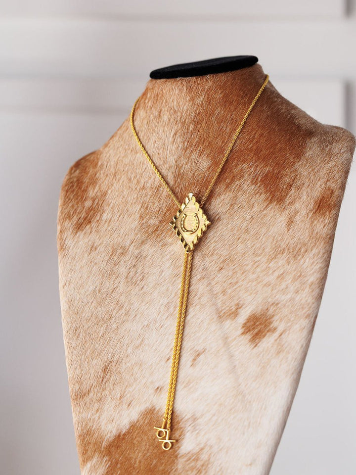 The Horseshoe Bolo necklace by Savannah Sevens western life{&}style features a gold-plated, rectangular engraved pendant with dangling charms, including a delicate horseshoe, displayed on a cowhidebust against a softly blurred background.