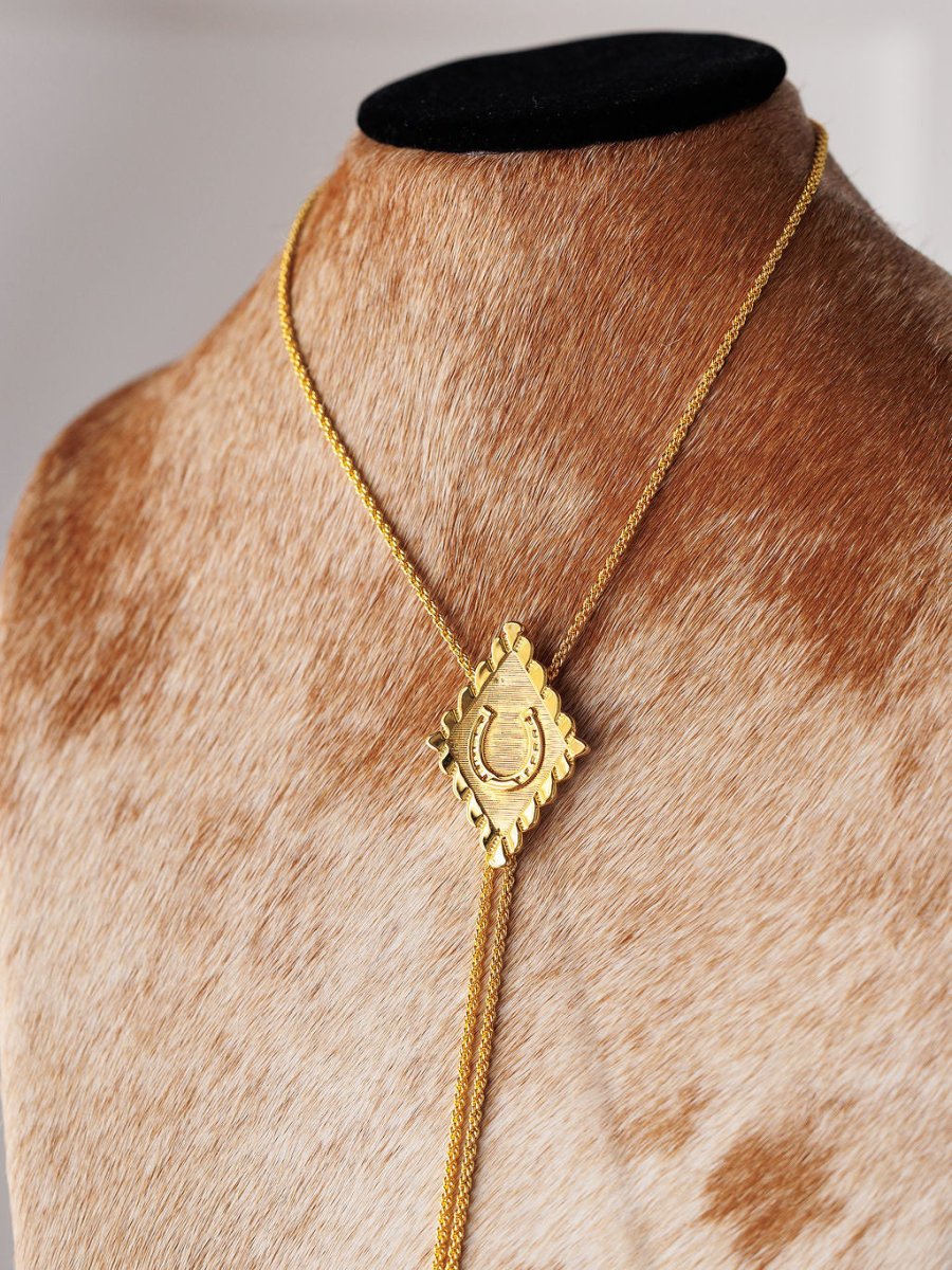 The Horseshoe Bolo by Savannah Sevens western life{&}style is a gold-plated necklace with a square, engraved pendant and fine chain strands, displayed on a brown and white cowhide mannequin bust.