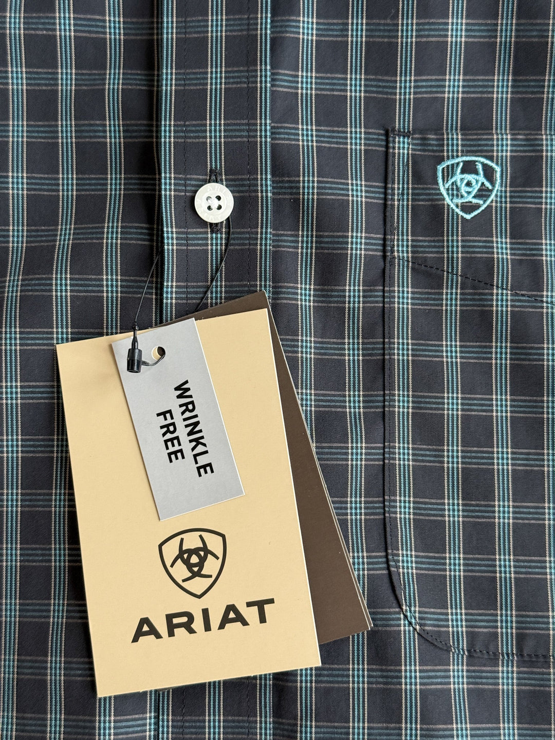 Close-up of a men’s plaid shirt in black and teal with a front pocket, featuring an embroidered Ariat logo and a beige tag reading ARIAT and WRINKLE FREE. This long sleeve button up offers both style and easy care with its wrinkle free fabric.