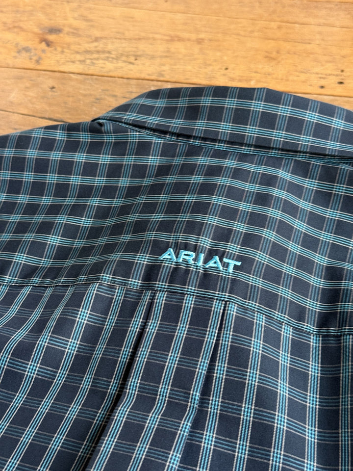 A close-up of the Houghson Ariat Men’s Plaid Button Up by Savannah Sevens western life{&}style, showcasing turquoise stitching and ARIAT embroidered on the back yoke. This wrinkle-free, long sleeve shirt is displayed lying on a wooden surface.
