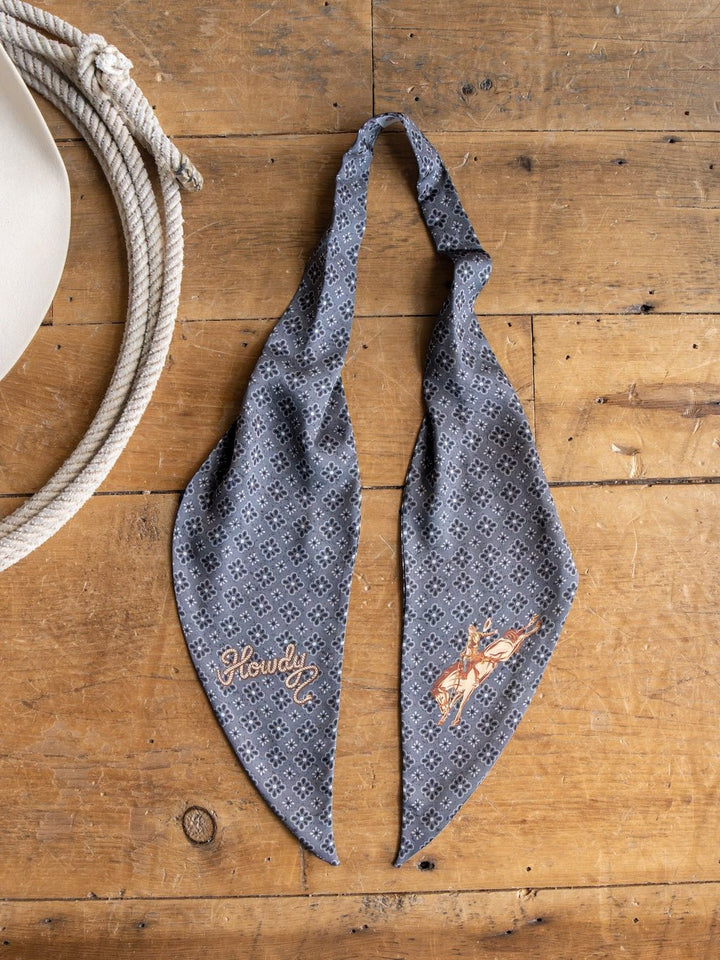 The Howdy Charcoal Satin Tie Scarf by Savannah Sevens western life{&}style, featuring a patterned gray design with "Howdy" and an embroidered rodeo rider, is laid flat on a wooden surface beside a coiled rope and a white cowboy hat.