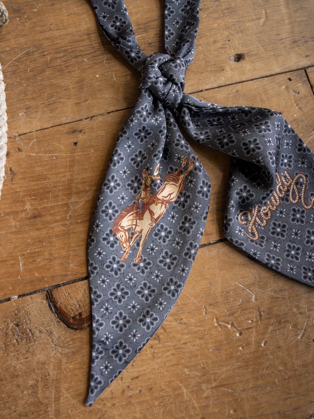 The Howdy Charcoal Satin Tie Scarf from Savannah Sevens western life{&}style, featuring a floral design on gray patterned poly chiffon, is displayed on a wooden surface with one end adorned by an illustration of a person riding a horse.