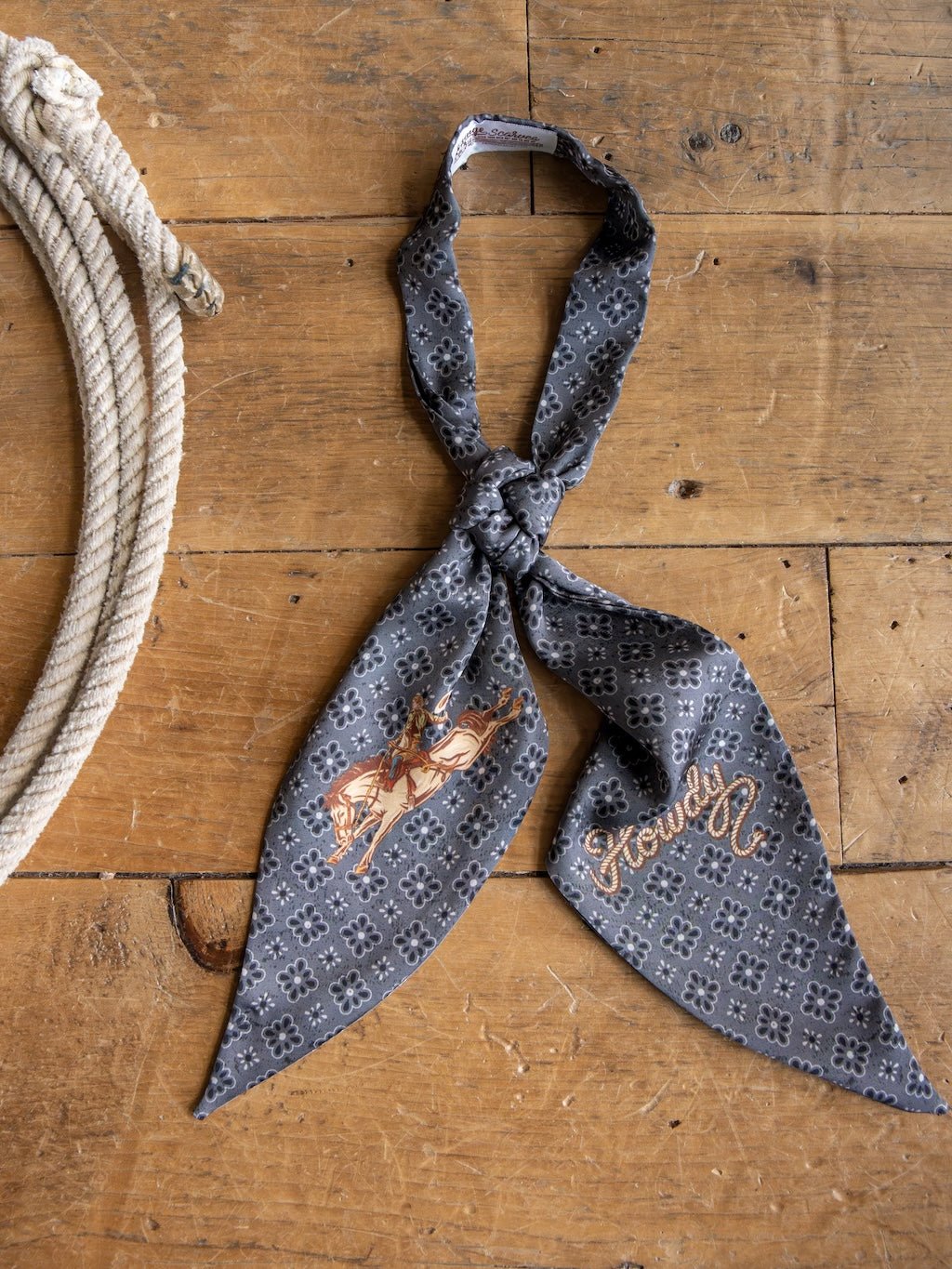 The Howdy Charcoal Satin Tie Scarf by Savannah Sevens western life{&}style, featuring a floral pattern, an illustrated cowboy on horseback, and the word "Howdy," is knotted on a wooden surface beside a coiled rope.