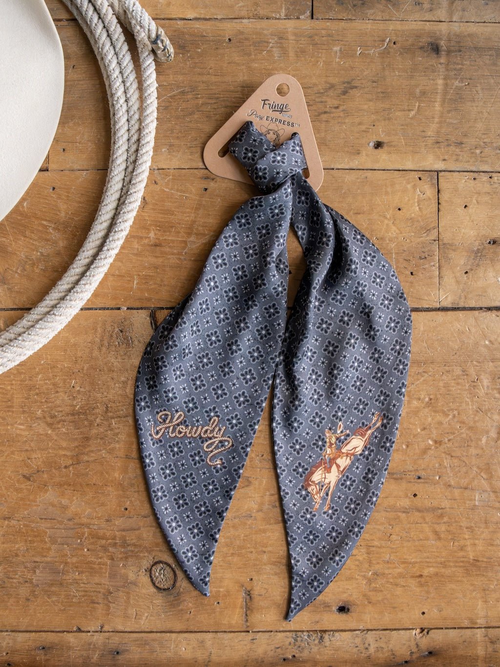 The Howdy Charcoal Satin Tie Scarf by Savannah Sevens western life{&}style is displayed on a wooden surface beside a coiled rope and the brim of a white hat. Featuring classic western motifs with "Howdy" and cowboy embroidery, this gray patterned accessory captures true western style and can also be styled as a concho pattern scarf for an extra touch of flair.