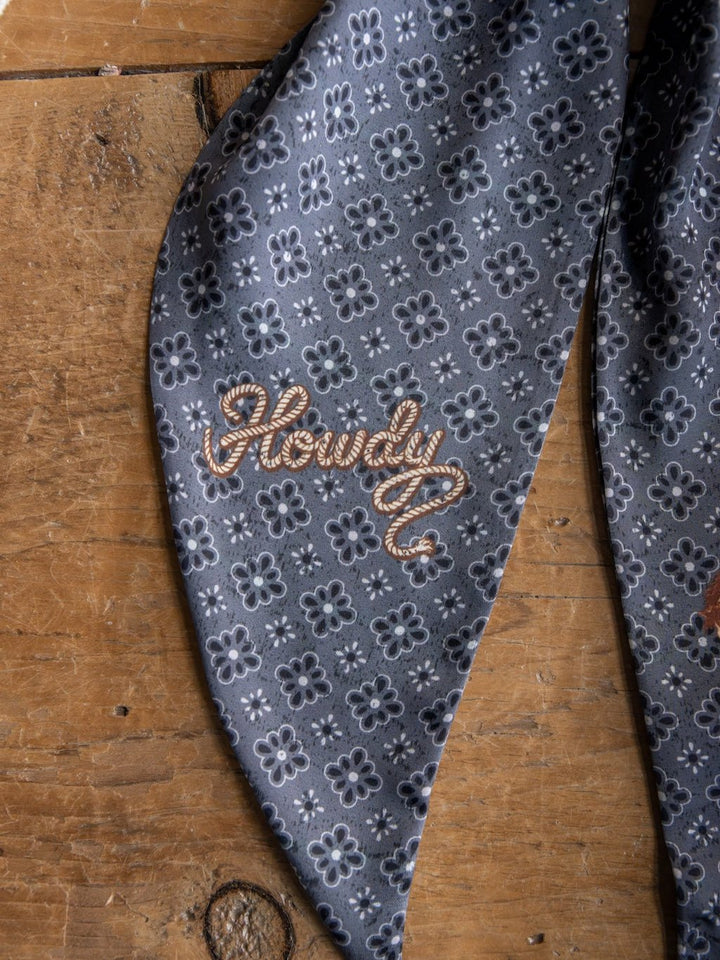 A charcoal patterned satin tie scarf from Savannah Sevens western life{&}style, featuring white floral designs and the word "Howdy" embroidered in tan thread, lying on a wooden surface.
