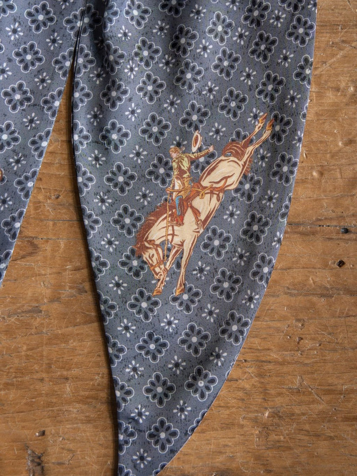 The Howdy Charcoal Satin Tie Scarf by Savannah Sevens western life{&}style features a gray patterned fabric with a cowboy riding a bucking horse, styled atop a wooden surface for true western flair.