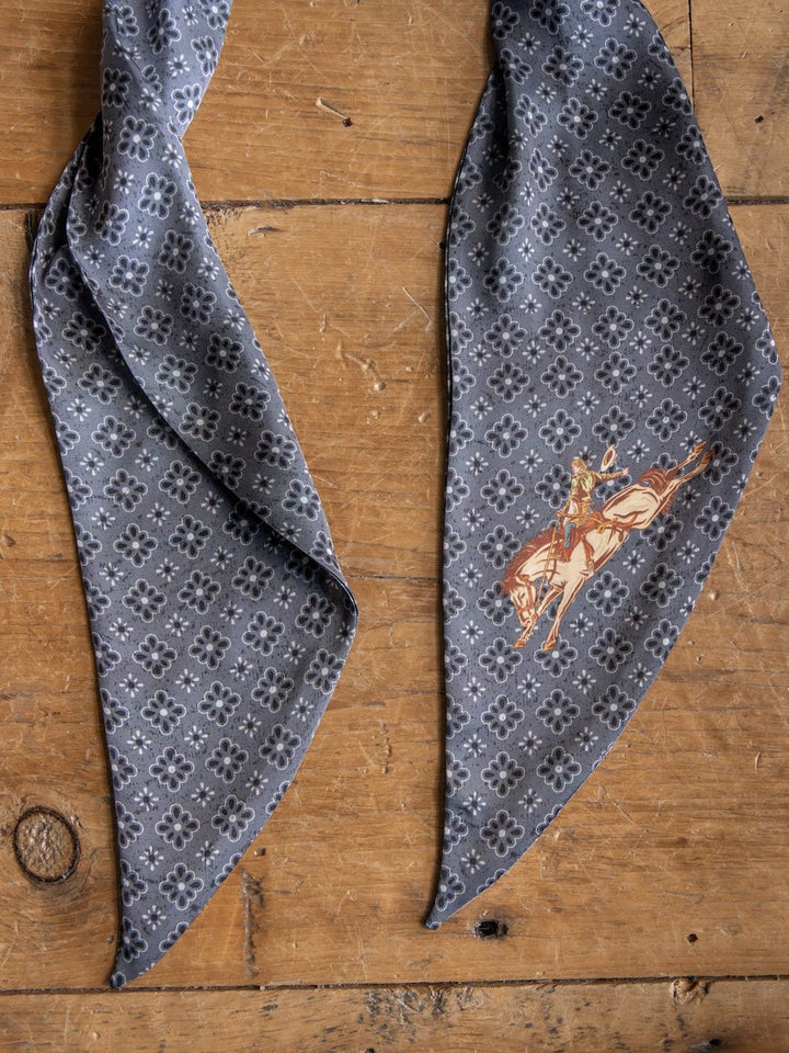 The Howdy Charcoal Satin Tie Scarf by Savannah Sevens western life{&}style, featuring black floral designs on a gray patterned poly chiffon fabric, rests on a wooden surface. One end of the scarf showcases an illustration of a cowboy riding a bucking horse.