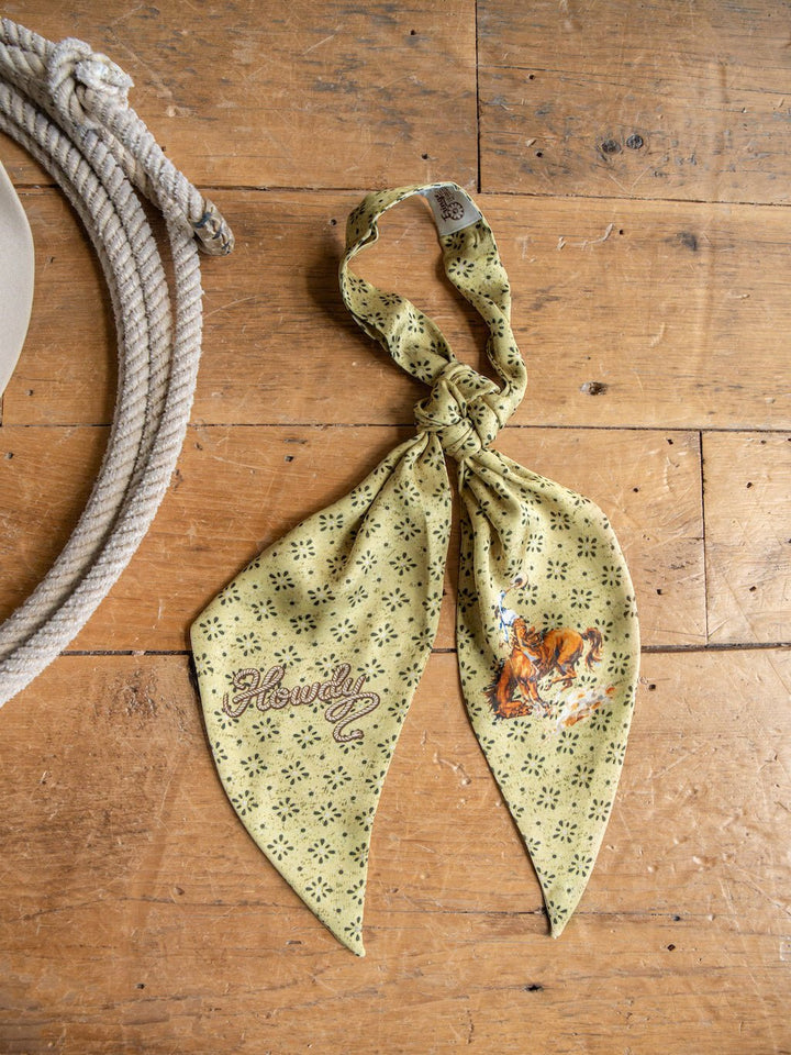 The Howdy Chartreuse Satin Tie Scarf by Savannah Sevens western life{&}style features a cowboy and horse illustration along with the word "Howdy" printed on vibrant yellow satin poly chiffon. Shown lying on a wooden floor near a coiled rope and the edge of a hat, this versatile accessory is perfect for styling as a western neck tie scarf.