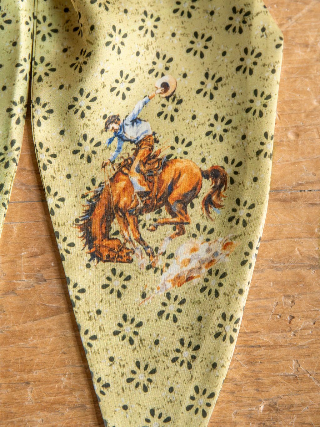 Featuring an illustrated cowboy riding a bucking horse with a raised hat against a wooden backdrop, the Howdy Chartreuse Satin Tie Scarf by Savannah Sevens western life{&}style showcases classic Western style with its yellow satin poly chiffon fabric adorned in a black floral pattern.