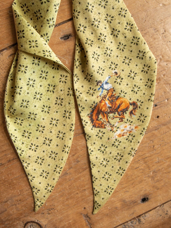 The Howdy Chartreuse Satin Tie Scarf by Savannah Sevens western life{&}style features a bold yellow pattern with black floral accents and a cowboy riding a bucking horse design, displayed on a wooden surface. Crafted from satin poly chiffon, this scarf boasts a light, smooth finish that enhances its unique western style.