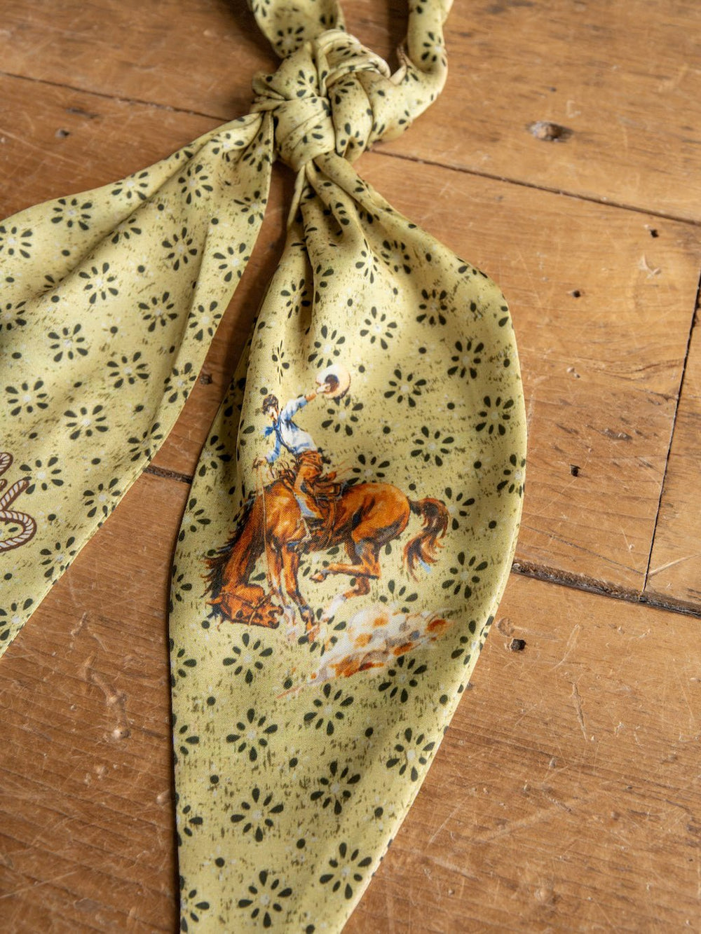 The Howdy Chartreuse Satin Tie Scarf by Savannah Sevens western life{&}style, featuring a yellow-green pattern with a cowboy riding a bucking horse, is displayed knotted on a wooden surface.