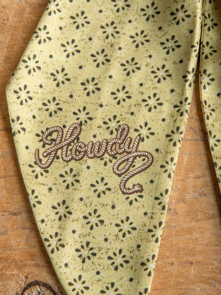 The Howdy Chartreuse Satin Tie Scarf by Savannah Sevens western life{&}style features a yellow-green satin poly chiffon fabric adorned with a small floral pattern and rope-style "Howdy" lettering, reminiscent of a western neck tie scarf and displayed on a wooden surface.