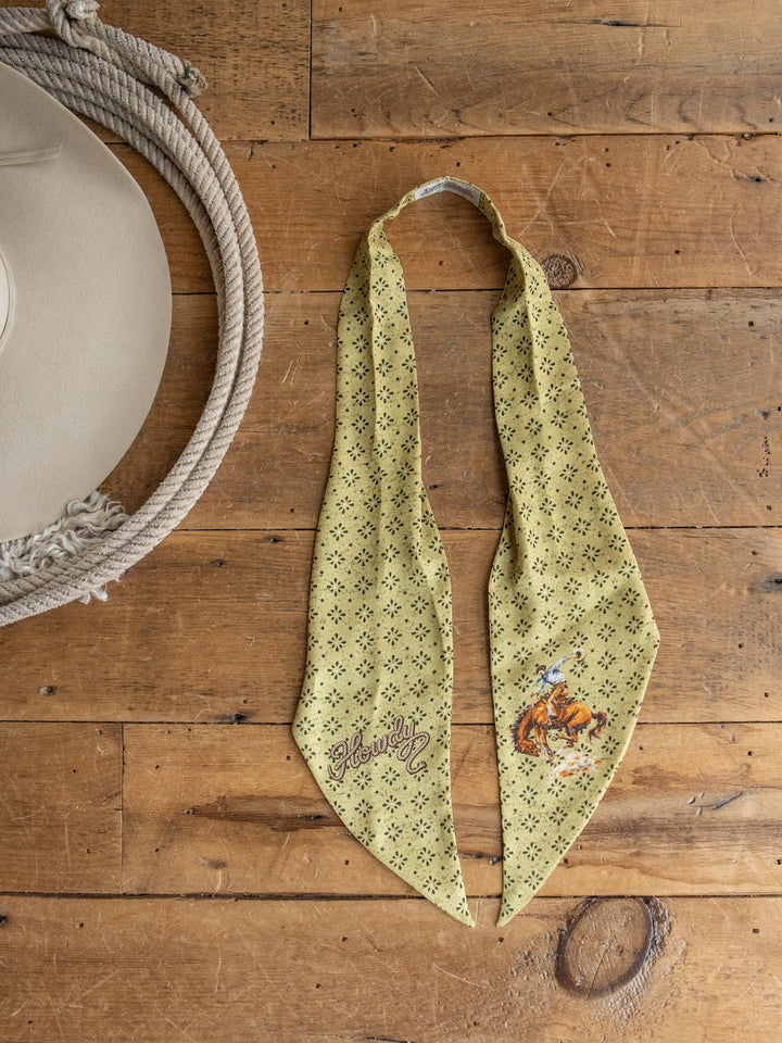 The Howdy Chartreuse Satin Tie Scarf from Savannah Sevens western life{&}style, featuring playful “Howdy” and cowboy graphics in a light green patterned satin, is displayed on a wooden surface alongside a beige cowboy hat and coiled rope.