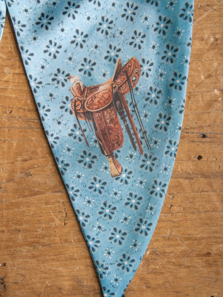 The Howdy Light Blue Satin Tie Scarf by Savannah Sevens western life{&}style features a blue satin fabric with a black floral pattern and a central illustration of a brown western saddle, displayed on a wooden surface.