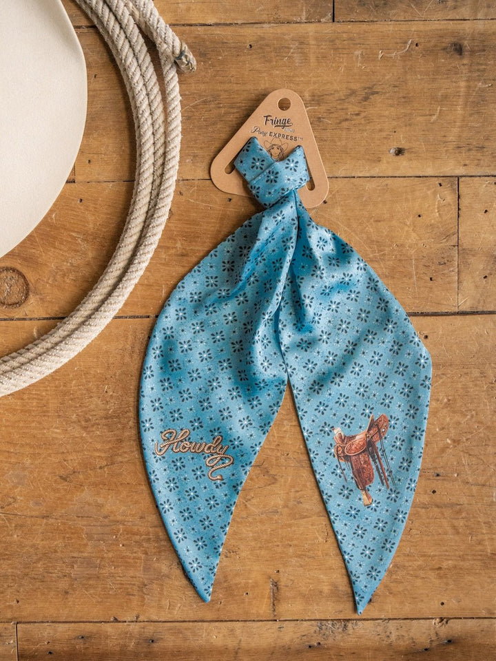 The Howdy Light Blue Satin Tie Scarf by Savannah Sevens western life{&}style features small floral patterns, "Howdy" text, and a saddle illustration. It is displayed on a wooden surface next to a coiled rope.
