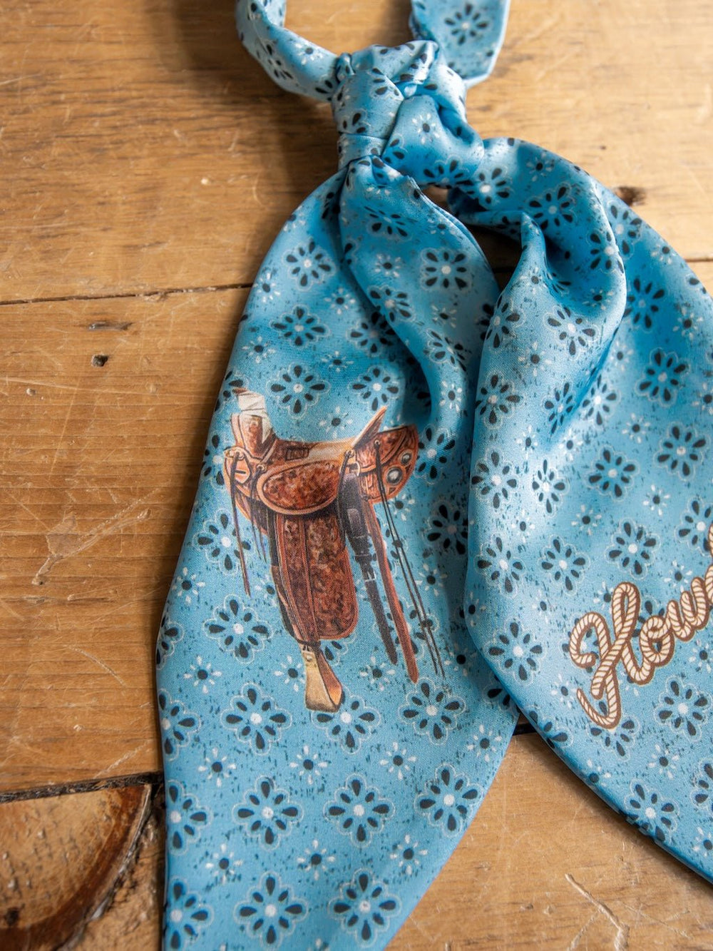 The Howdy Light Blue Satin Tie Scarf by Savannah Sevens western life{&}style, featuring a brown saddle print and the word "Howdy" in script, is displayed on a wooden surface.