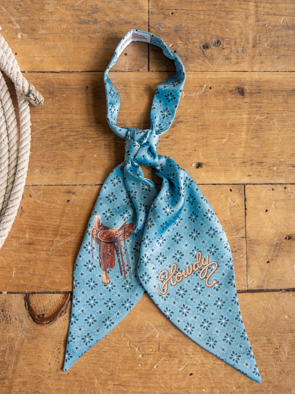A Howdy Light Blue Satin Tie Scarf from Savannah Sevens western life{&}style, showcasing a blue patterned design with a saddle illustration and “Howdy” embroidery, is laid on a wooden surface beside a coiled rope.