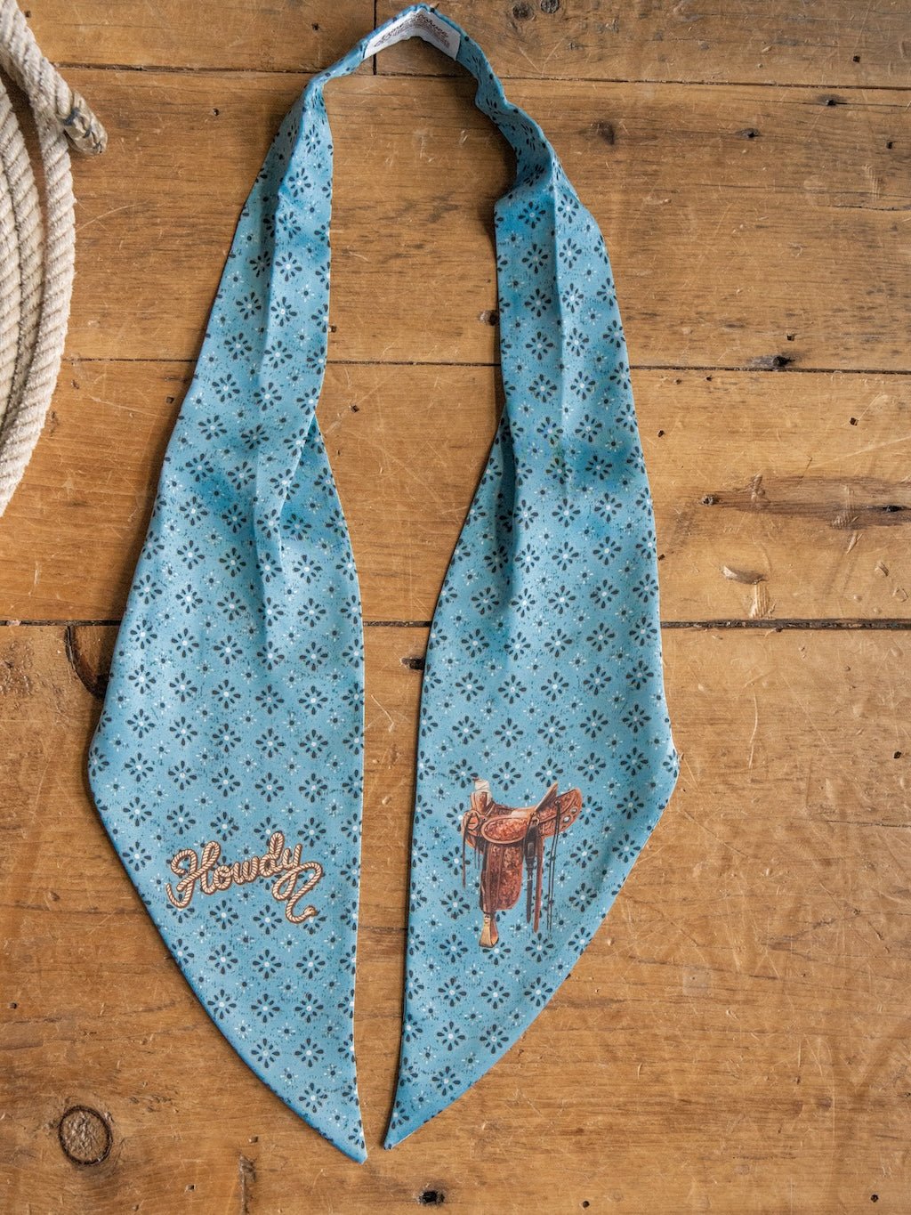 A Howdy Light Blue Satin Tie Scarf by Savannah Sevens western life{&}style, adorned with small black patterns and embroidered with "Howdy" and a saddle, is laid flat on a wooden surface beside a coiled rope.