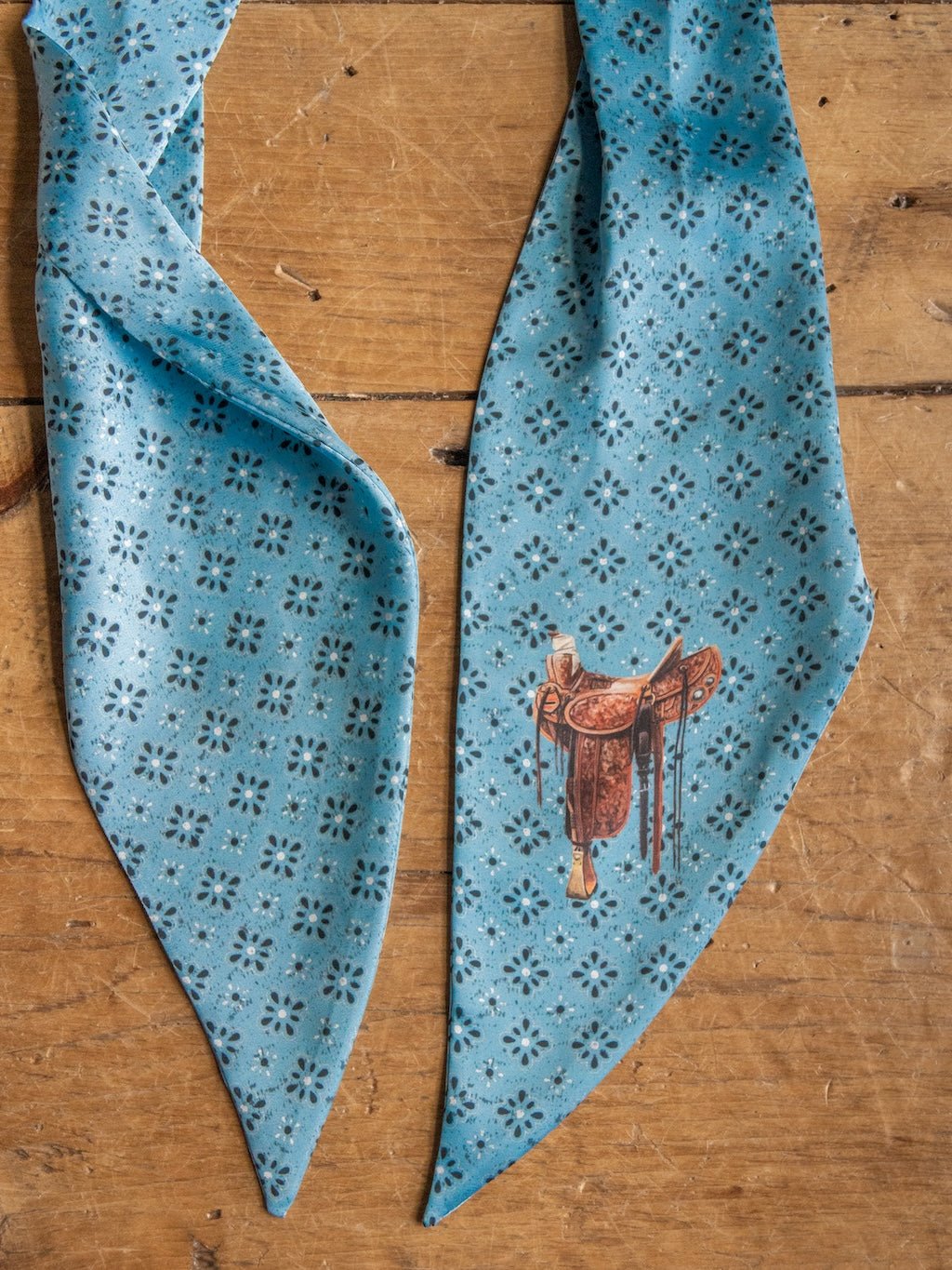 The Howdy Light Blue Satin Tie Scarf by Savannah Sevens western life{&}style, featuring an illustration of a brown saddle and subtle concho pattern, rests on a wooden surface.