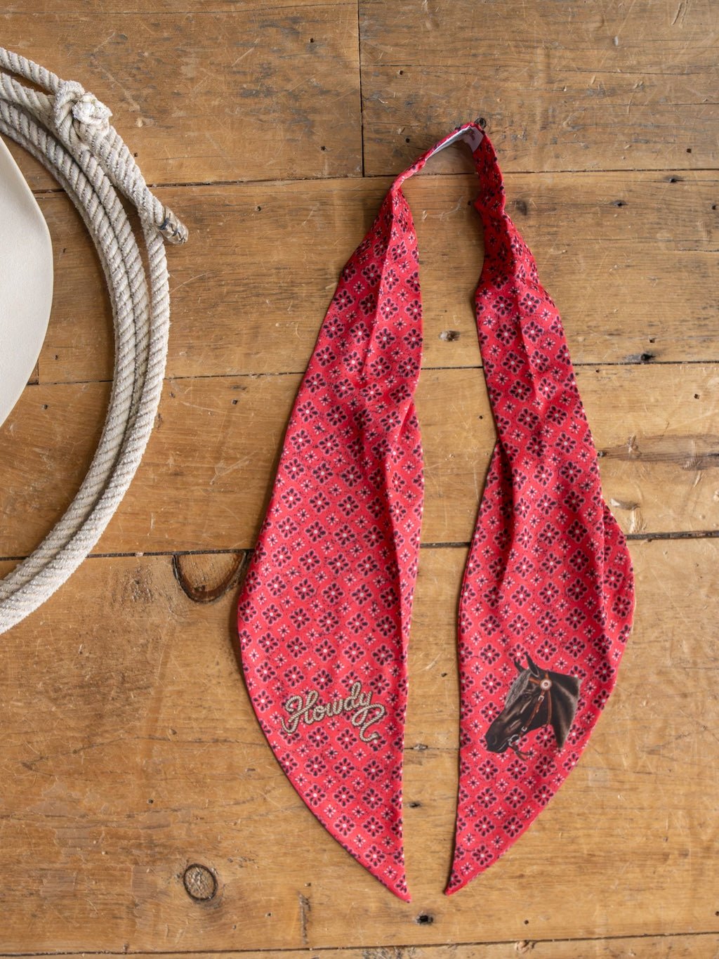 The Savannah Sevens western life{&}style Howdy Red Satin Tie Scarf, featuring a "Howdy" greeting and a horse head design, is displayed on a wooden surface beside a coiled rope and a tan cowboy hat.