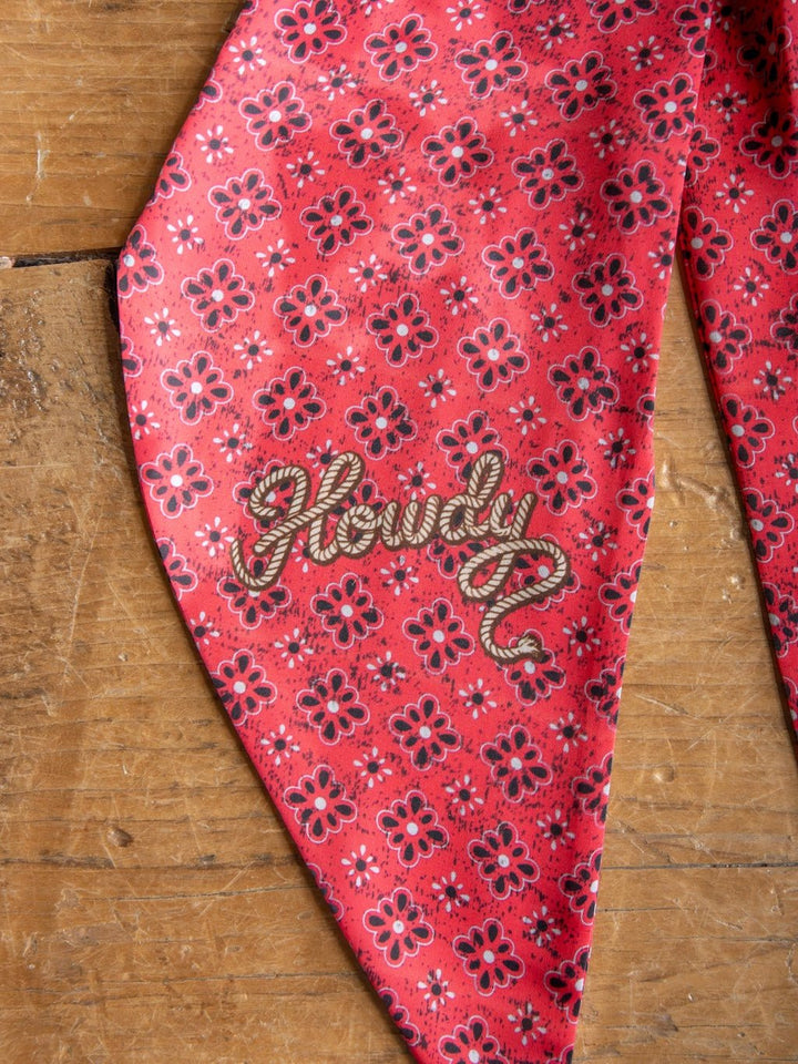 A red satin tie scarf from Savannah Sevens western life{&}style, featuring a floral bandana pattern, rests on a wooden surface. "Howdy" is embroidered in rope-like letters on one corner, making it the perfect western neck accessory.