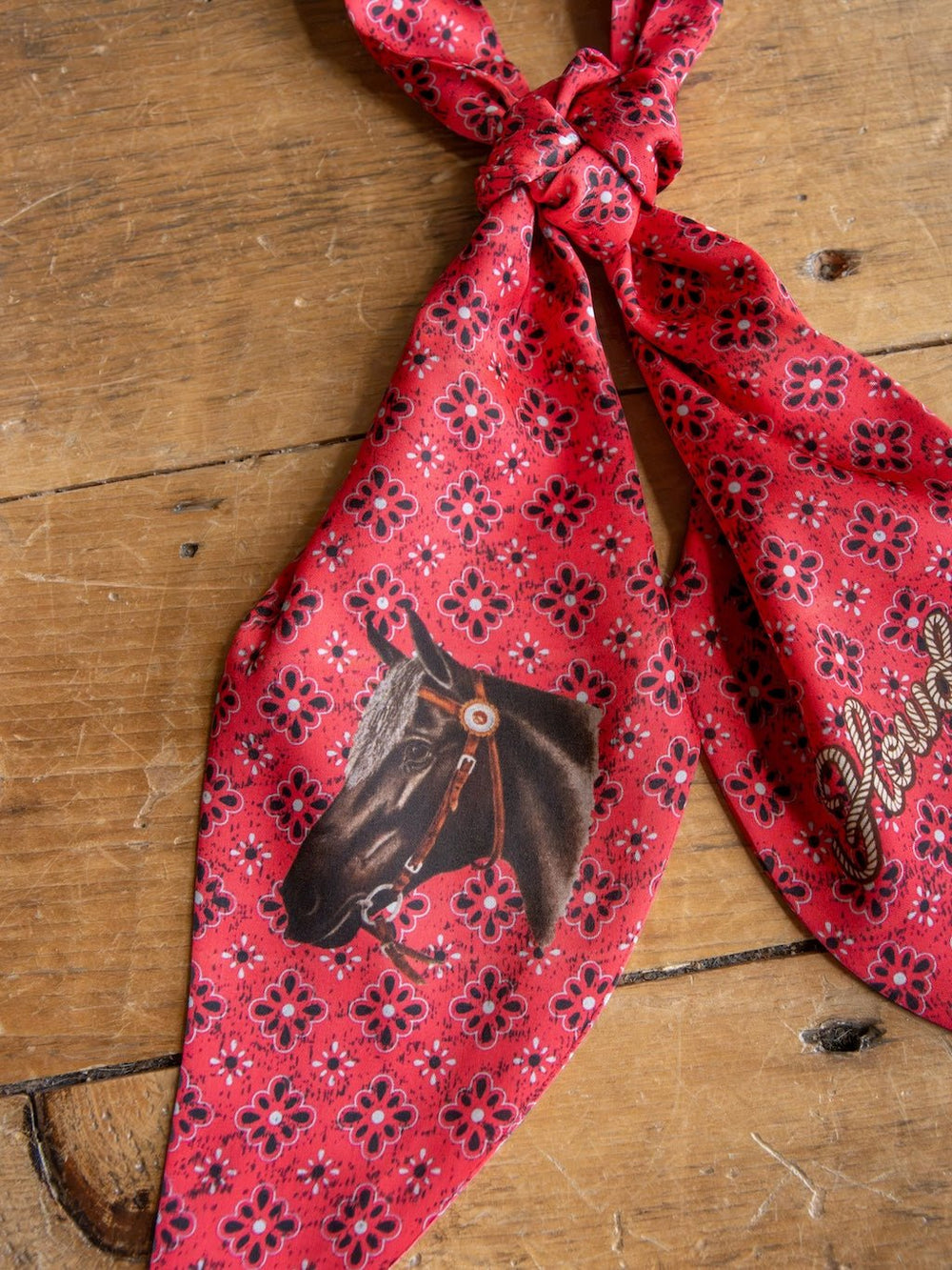 The Howdy Red Satin Tie Scarf from Savannah Sevens western life&style, featuring a floral pattern and a printed horse head with a bridle, is knotted on a wooden surface. This western neck scarf brings classic style to any outfit.